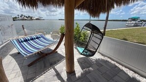 A hammock and swing chair under a thatched roof, overlooking a body of water with a boat in the distance. - Waterfront Matlacha views! 2/2 Heated Pool Home (Matlacha)