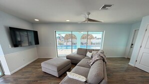 A living room with a sectional sofa, ottoman, and a flat-screen TV mounted on the wall. - Waterfront Matlacha views! 2/2 Heated Pool Home (Matlacha)