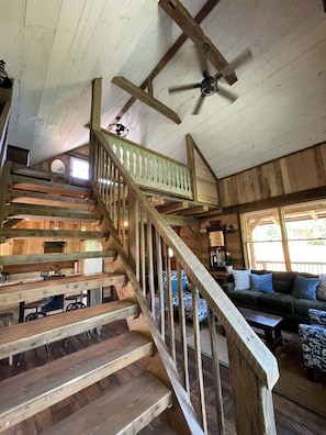 Eine Holztreppe mit Geländer, ein Deckenventilator und eine Couch in einem blockhausartigen Innenraum.