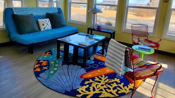 A living room with a blue sofa, a coffee table, a patterned rug, and a chair with a striped cushion. - Mermaid's Mini Mansion (Eastport)