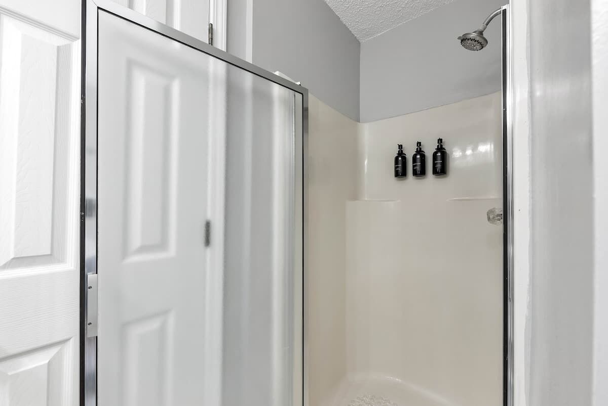 A white door with a metallic handle, a shower area with a handheld showerhead, and three bottles mounted on the wall.