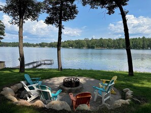 A lakeside outdoor seating area with Adirondack chairs, a fire pit, and a dock extending into the water. - (Nisswa)