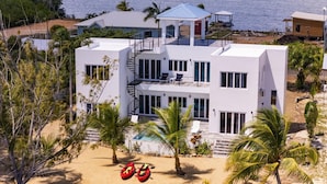 Een modern huis met twee verdiepingen, een zwembad, omringd door palmbomen en een strand.