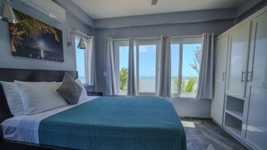Een slaapkamer met een bed, een nachtkastje, een raam met gordijnen, een plafondventilator en uitzicht op de oceaan.