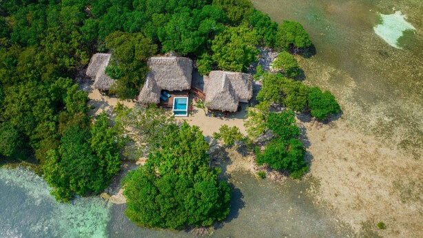 Vista aérea de una isla tropical con cabañas de techo de paja, una piscina y abundante vegetación.