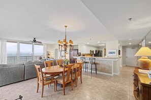 A spacious living area with a dining table, sofa, kitchen, and a balcony with a view. - Emerald Coast Escape – End Unit Steps from the Sand (Pensacola Beach)