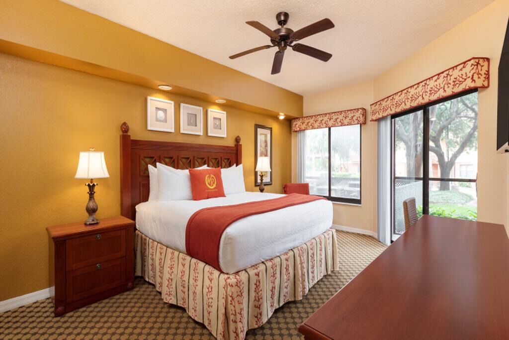 A hotel room with a bed, a desk, a ceiling fan, and large windows.
