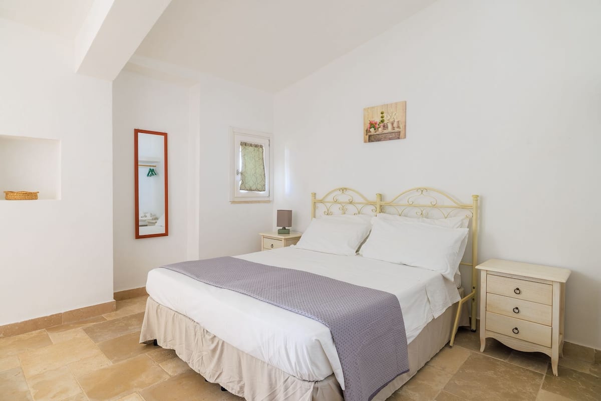 Quarto com cama, mesas de cabeceira, cómoda, janela com cortinas e quadro na parede.