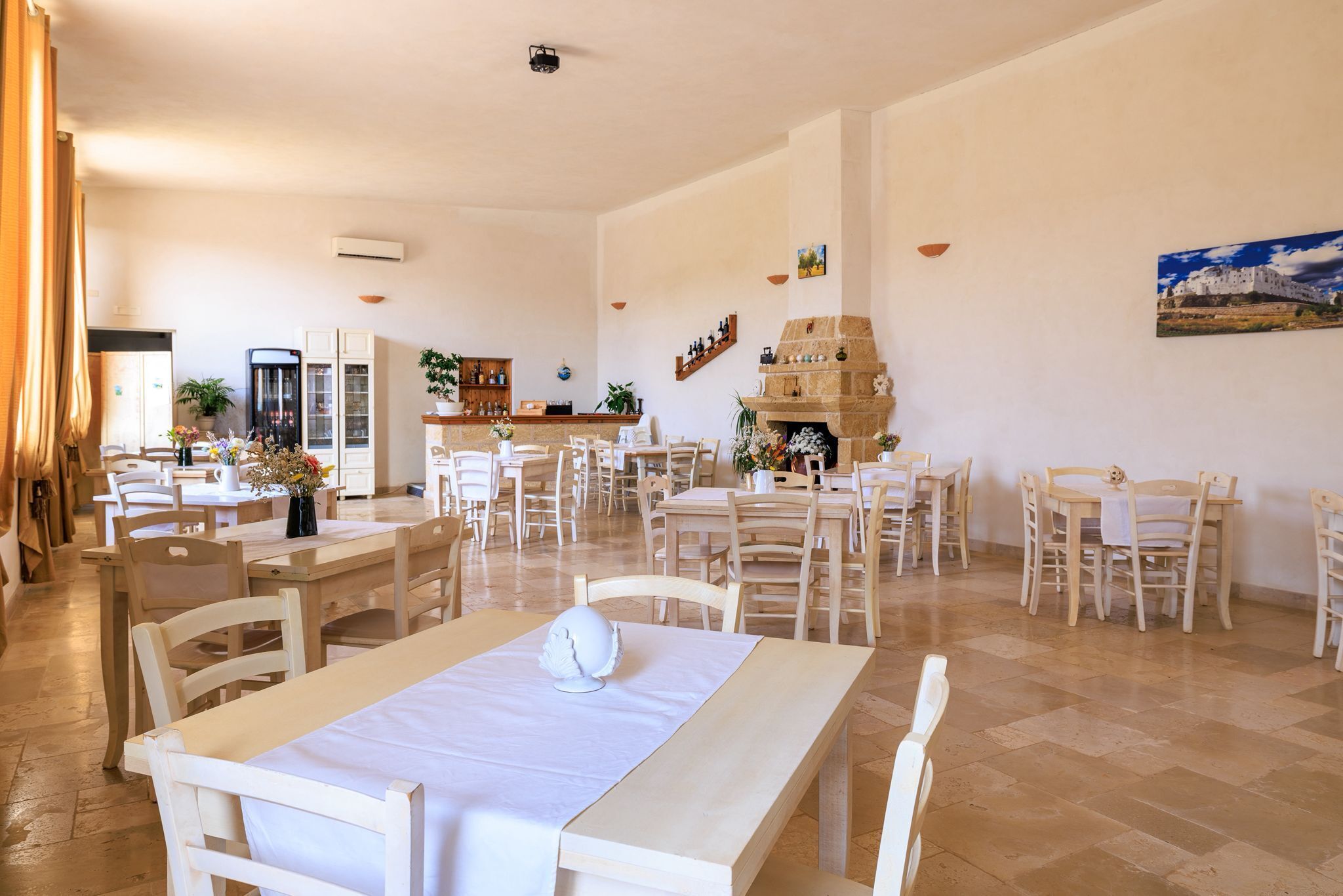 A spacious dining area with multiple tables and chairs, a stone fireplace, and a view of the sea through the windows.
