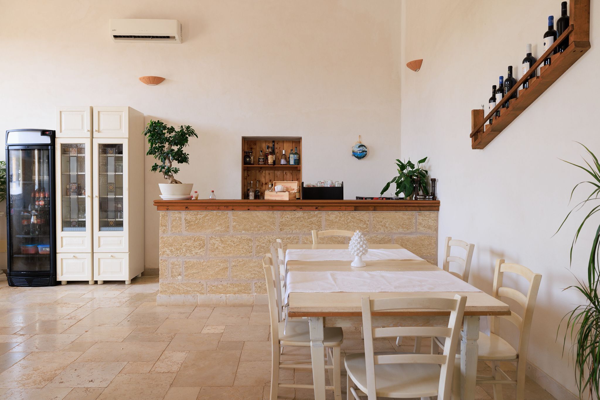 Un comedor con mostrador de piedra, sillas blancas, una mesa con mantel blanco, una planta en maceta y una estantería para vinos en la pared.