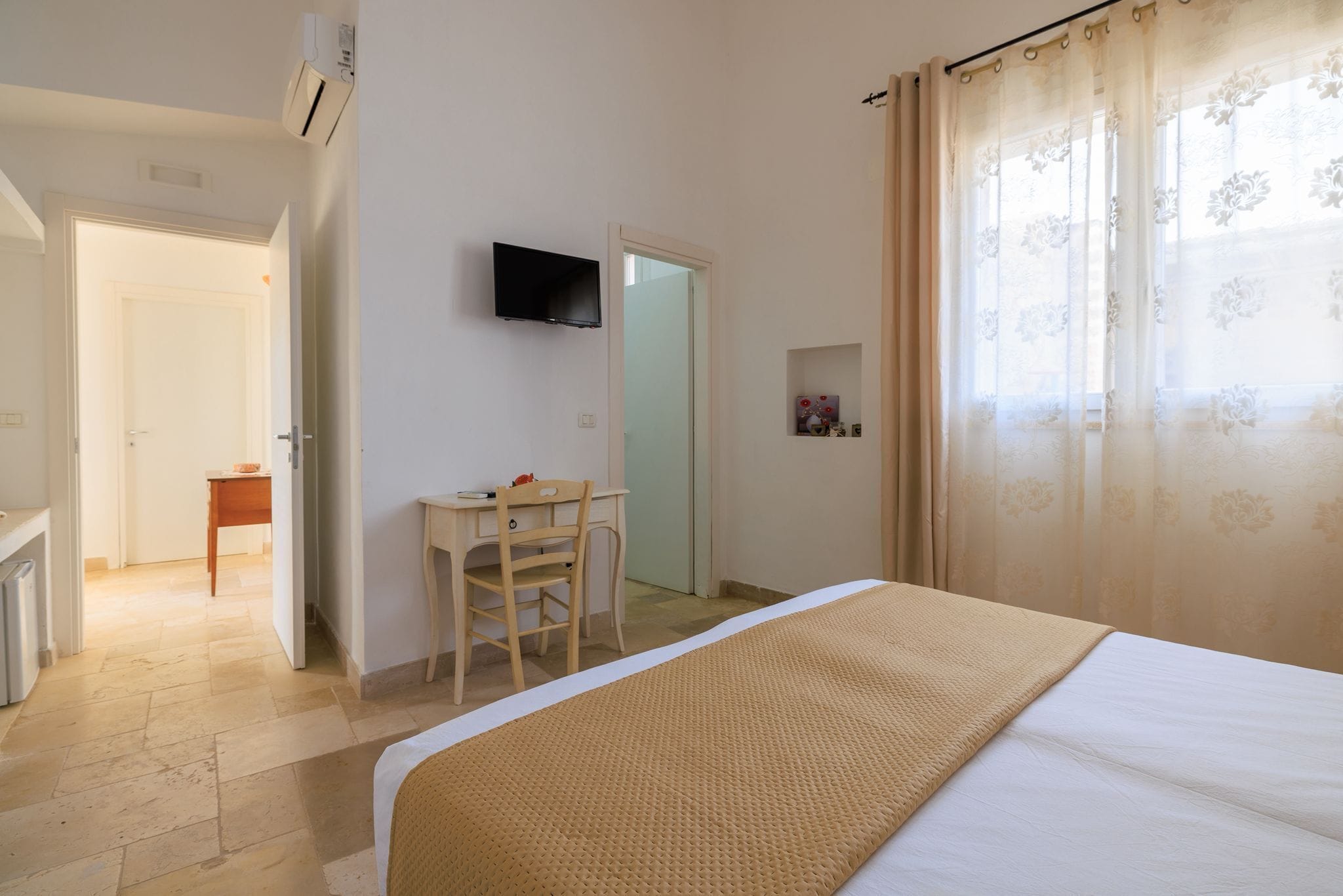 Un dormitorio con una cama, un escritorio con silla, un televisor en la pared y una puerta que da a otra habitación.