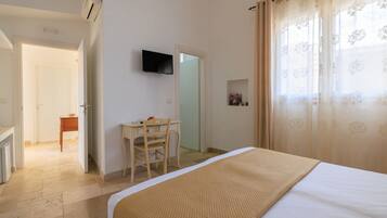 Un dormitorio con una cama, un escritorio con silla, un televisor en la pared y una puerta que da a otra habitación.