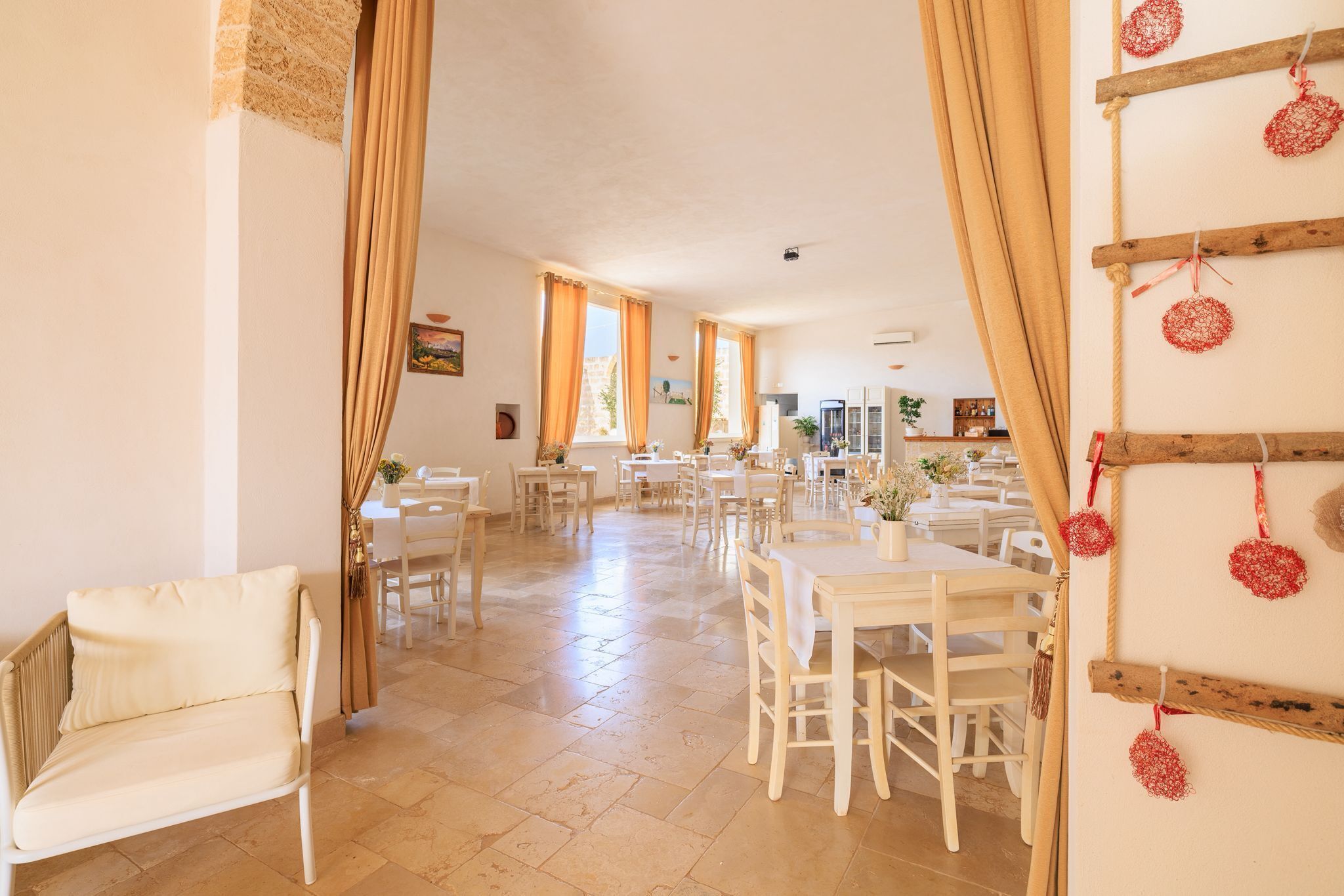 A spacious dining area with white tables and chairs, large windows with curtains, and a wooden wall decoration.
