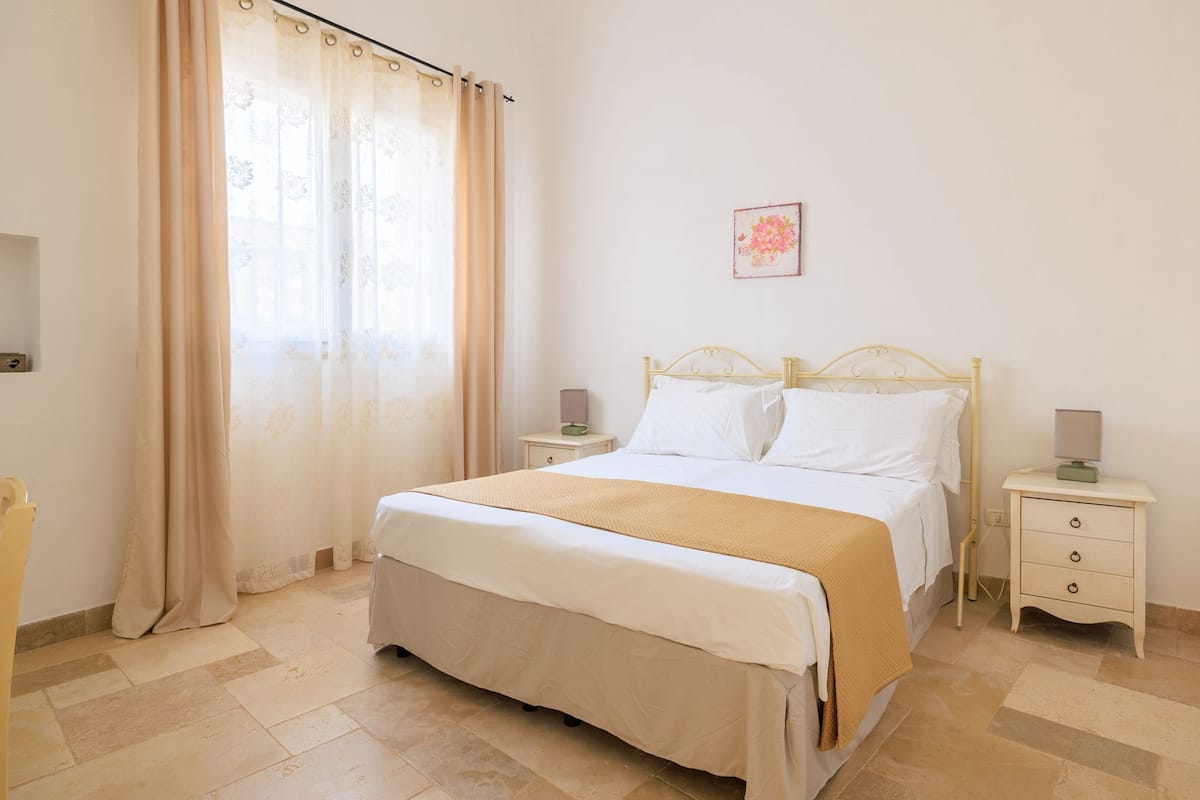 Quarto com cama, cortinas, mesa de cabeceira e um quadro na parede.