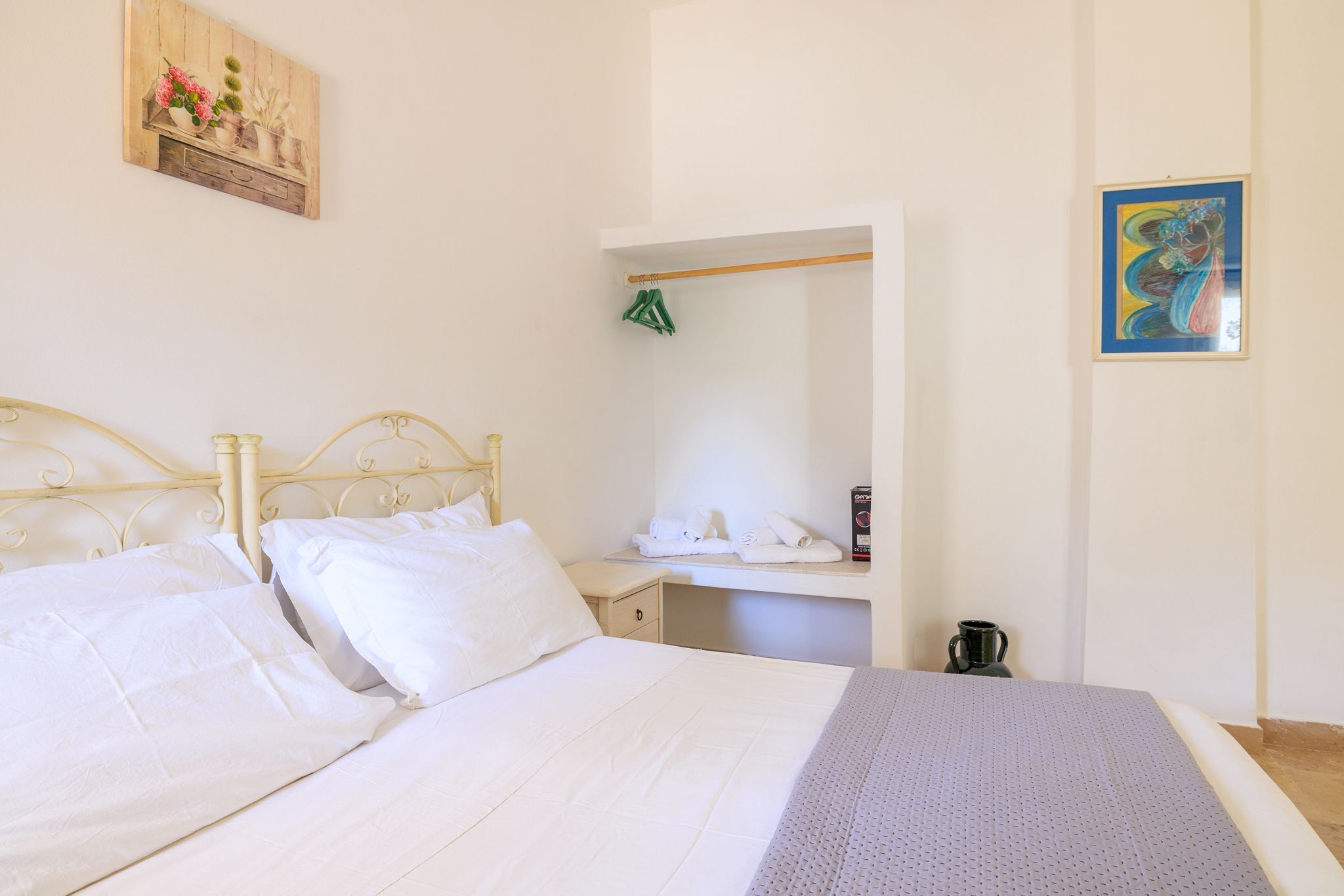 A neatly made bed with white linens, a small bedside table with a cup, and a wall-mounted shelf with folded towels.