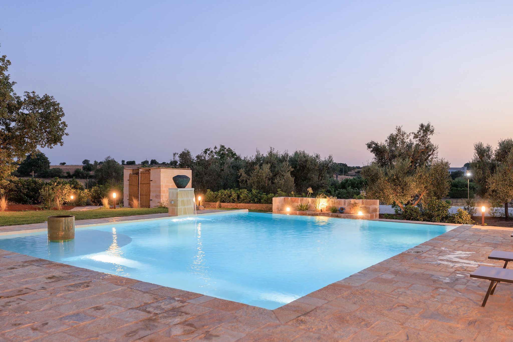 A rectangular swimming pool with a stone border, surrounded by a landscaped garden with trees and shrubs, and a stone building with a circular window in the background.