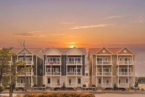 Una hilera de edificios de varios pisos con balcones, una bandera y un atardecer al fondo.