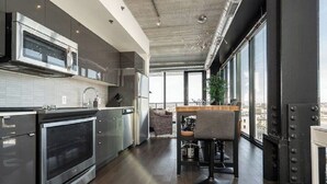 A modern kitchen with stainless steel appliances, a dining area with a high table and chairs, and large windows offering a city view.