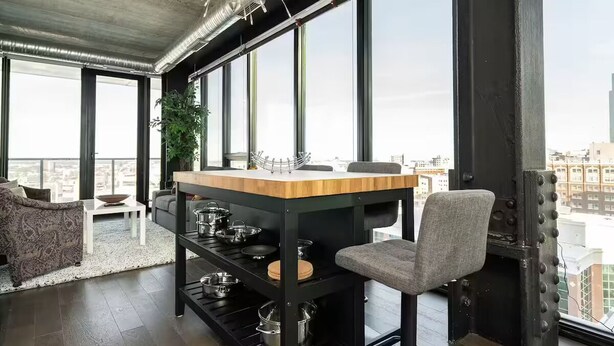 A modern kitchen with a wooden countertop, metal shelves, and a view of the city through large windows.
