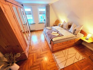A bedroom with a wooden bed, a herringbone floor, a window with curtains, and a potted plant. - Make your time out special - we look forward to welcoming you. (Zingst)