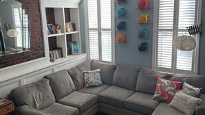 A living room with a sectional sofa, a small stool, a bookshelf, and a wall-mounted shelf with colorful items. - Marvelous 3-bedroom townhome in vibrant upper Lawrenceville (Pittsburgh)