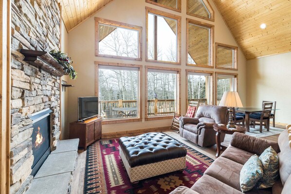 A cozy living room with a stone fireplace, large windows, a comfortable sofa, and a television. - Aspen Grove Retreat: Sweeping Views, Fireplace (Angel Fire)