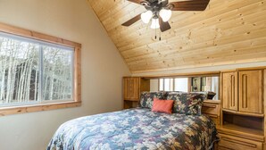 Un dormitorio con una cama grande, cabecera de madera, estanterías empotradas, ventilador de techo y ventana con vistas a árboles.