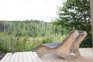 En träbänk med ett böjt ryggstöd placerad på ett trädäck med utsikt över en skogbeklädd sluttning.