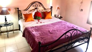 Una cama con una colcha morada, una almohada blanca y dos almohadas naranjas. Hay una lámpara sobre una mesita de noche junto a la cama.
