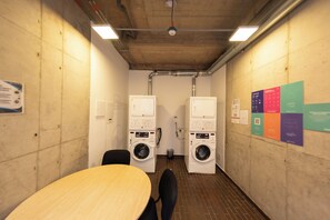 Een wasserette met twee wasmachines, een tafel, stoelen en informele posters aan de muur.