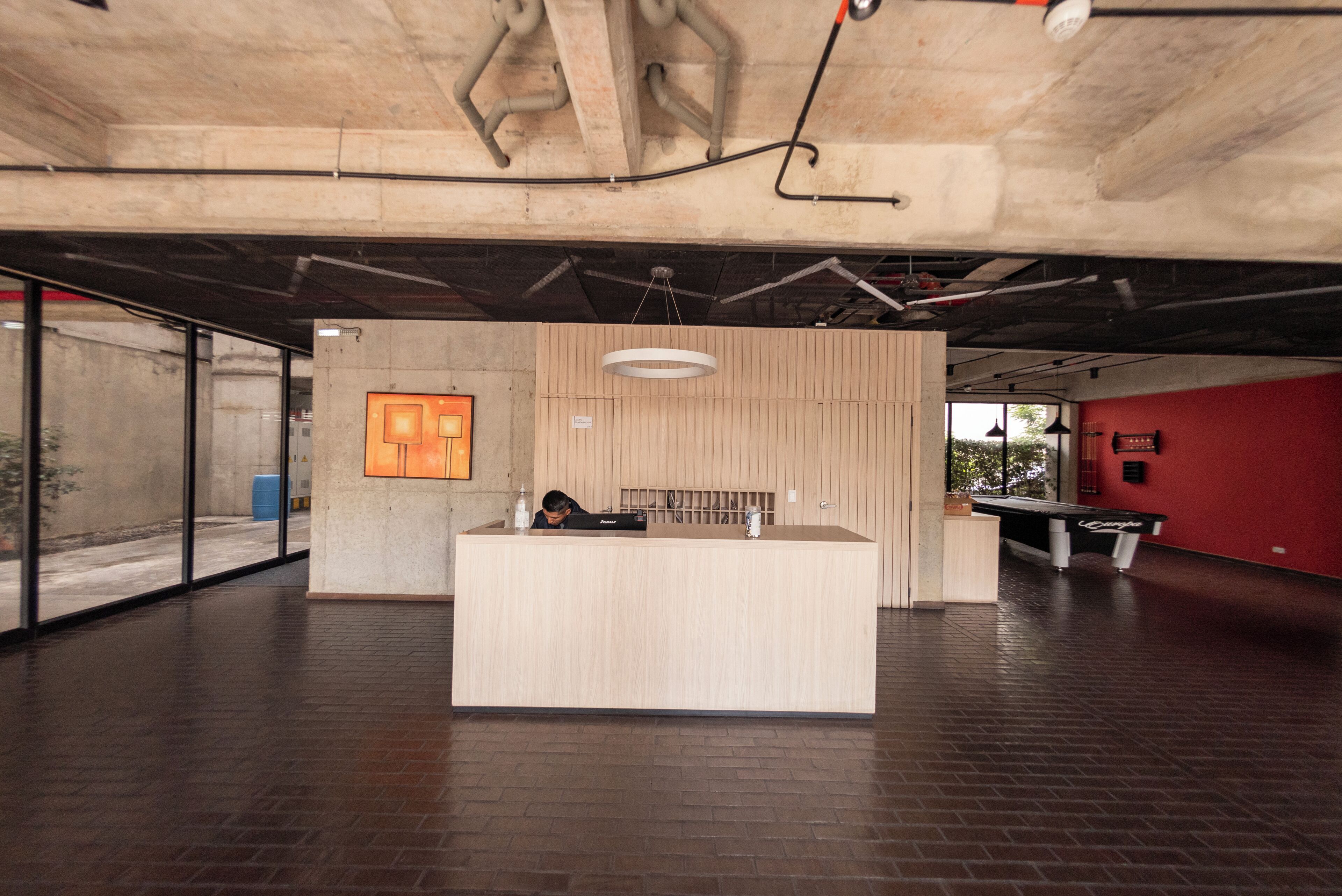 A reception desk in a modern building with a concrete ceiling, a wooden reception desk, a hanging light fixture, and a billiards table.