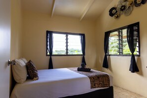 A hotel room with a bed, two windows with curtains, a ceiling fan, and a decorative wall piece. - Hidden Haven—Private Mountain View - Retreat in Nature (Rarotonga)