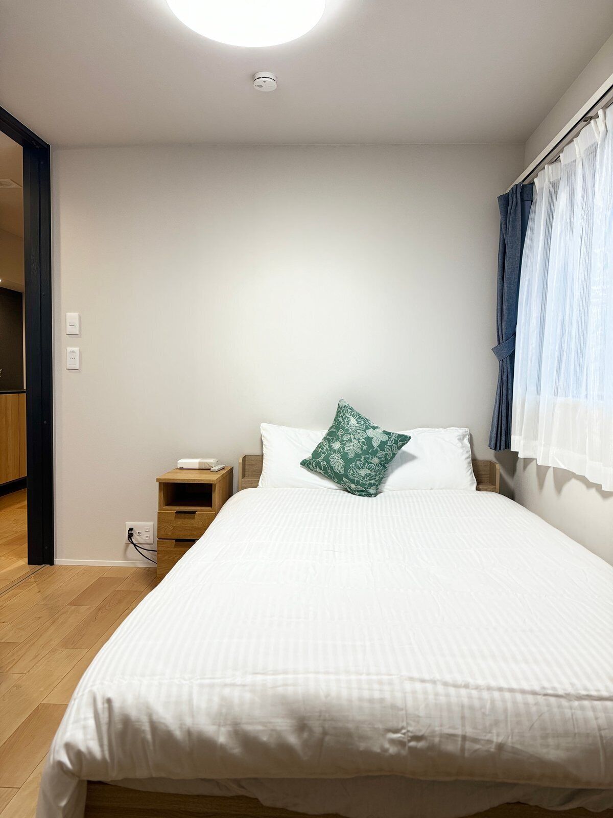 Un dormitorio con una cama, una mesita de noche, un enchufe y una ventana con cortinas.