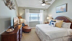 Ein Schlafzimmer mit einem großen Bett, einem Deckenventilator, Blick aufs Meer, einem hölzernen Kopfteil und einem Schreibtisch mit Lampe.