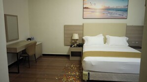 Una habitación de hotel con cama, escritorio, silla, mesita de noche, lámpara y un cuadro con una escena de playa.