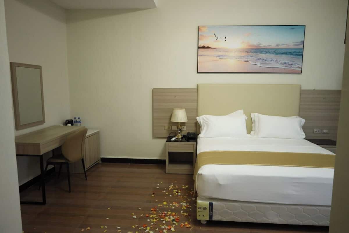 Ein Hotelzimmer mit Bett, Schreibtisch, Stuhl, Nachttisch, Lampe und einem Bild mit Strandmotiv.