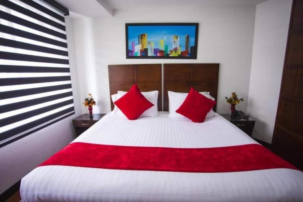 Ein Doppelbett mit weißer und roter Bettwäsche, darüber ein Stadtbildgemälde und ein Fenster mit schwarz-weißen Jalousien.