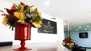 Een receptiegedeelte met een rode vaas bloemen, een zwart bord met 'Bogotá Real' en een kroonlogo, en een televisie aan de muur.