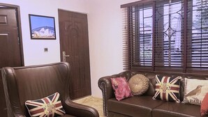 Ein Wohnzimmer mit braunem Ledersessel, braunem Sofa mit Zierkissen, einem Fenster mit Jalousien und einem gerahmten Bild an der Wand.