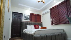 Ein Schlafzimmer mit Bett, Tür, Fenster mit Jalousien und Deckenleuchte.