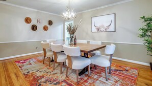 Un comedor con una mesa de madera, sillas blancas, una araña y un tapete estampado.
