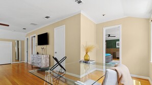 Una habitación amplia con una mesa de vidrio, piso de madera, una televisión de pantalla plana y una puerta que da a otra habitación.
