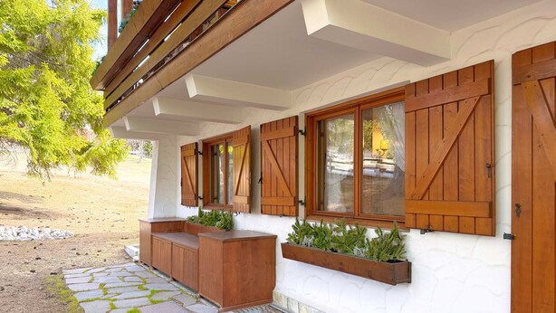 Un espacio al aire libre cubierto con suelo de piedra, bancos de madera y macetas, todo ello frente a una pared blanca con contraventanas de madera.