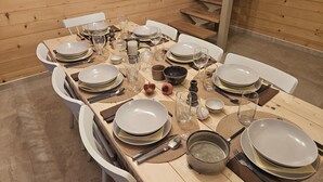 Una mesa de comedor dispuesta con platos, tazones, vasos y cubiertos, rodeada de sillas blancas en un ambiente de madera.