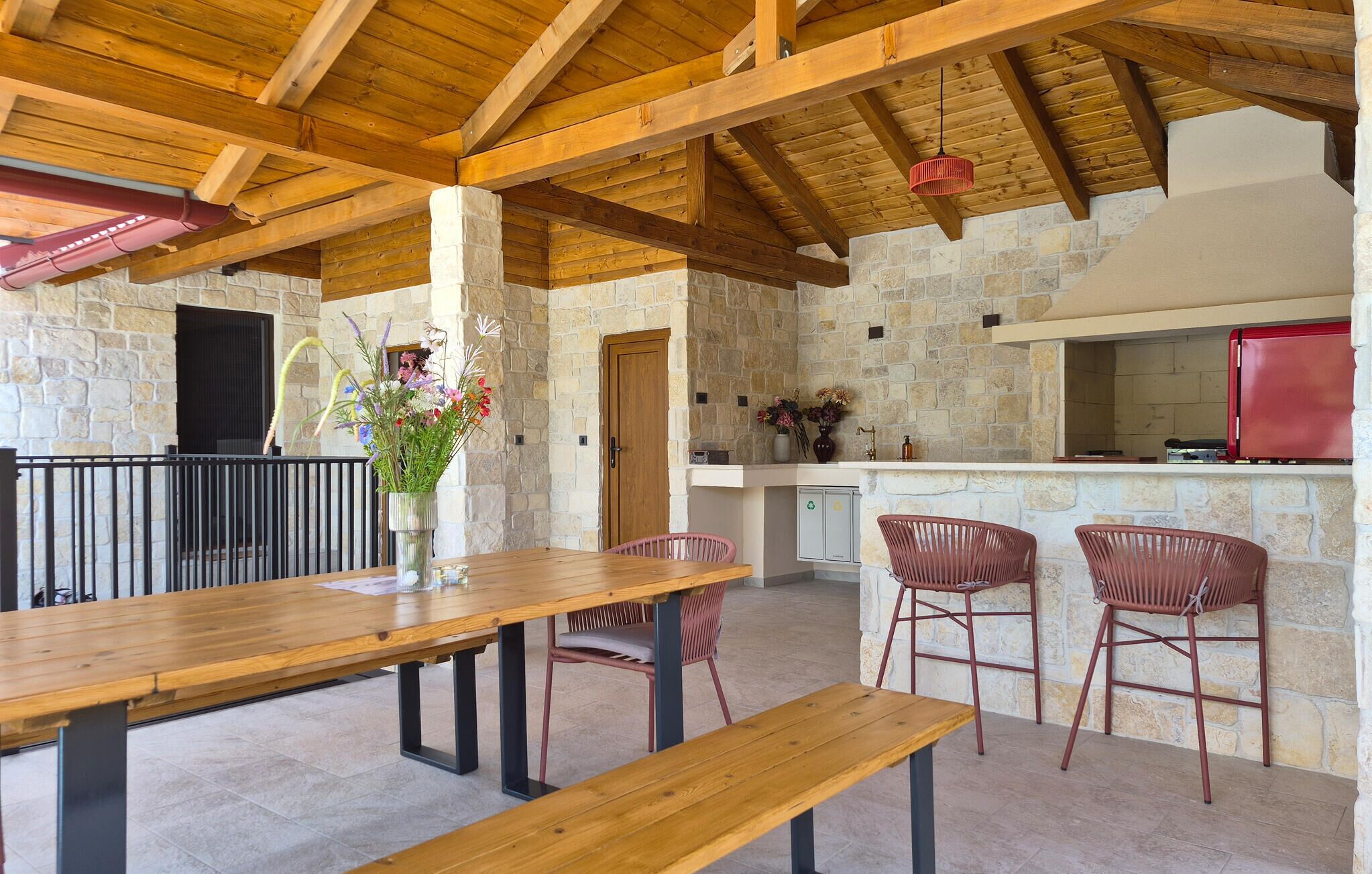 An outdoor dining area with a wooden table and benches, stone walls, and a stone fireplace.