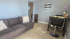 A modern living room with a grey sofa, two white cushions, a small black chair, a wooden side table, and a wall-mounted shelf. - #15 Guest house Midtown stadium B (Tampa)