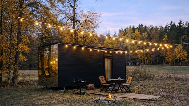 Eine kleine Holzhütte mit Tisch und Stühlen vor einer baumbestandenen Umgebung, beleuchtet mit Lichterketten.