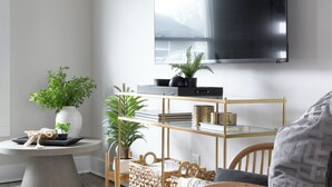 Ein modernes Wohnzimmer mit Flachbildfernseher, rundem Couchtisch, Beistelltisch aus Weidengeflecht, Korbbasket und Holzstuhl, sowie einem Blumentopf.
