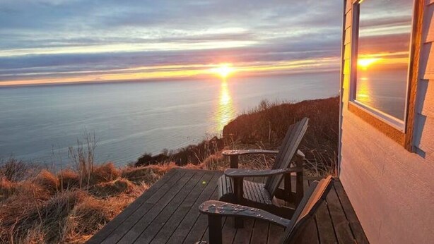 A wooden deck with two Adirondack chairs overlooking a sunset over a body of water.