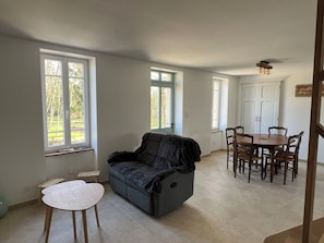 Una habitación con sofá, una pequeña mesa redonda y una mesa de comedor con sillas. Hay ventanas con vistas a zonas verdes.